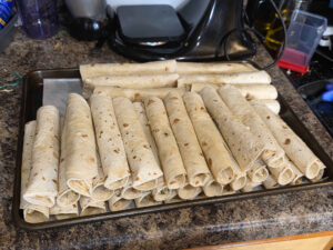 A cookie sheet with assembled taquitos stacked on it. There are almost sixty.